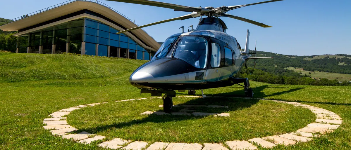 A silver helicopter on a grass helipad under a blue sky